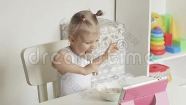 一个小女孩坐在孩子们`桌子旁，用勺子吃粥，看着电子药片的屏幕